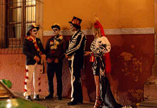 A woman and three men stand beneath a glowing street lamp in Dia de Muertos costumes with skull-painted faces.