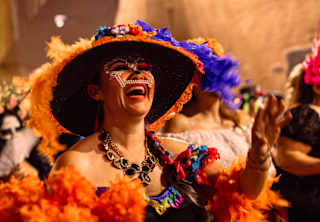 A woman in a flamboyant skirt and hat in pinks and purples, with marigold-orange feathers, enjoys the Dia de Muertos parade.