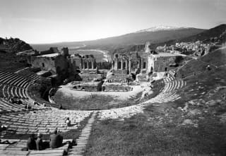 Monochrome image of Teatro Antico di Taormina with arced stone steps and backdrop of ruins, coastline and snow-capped Etna.