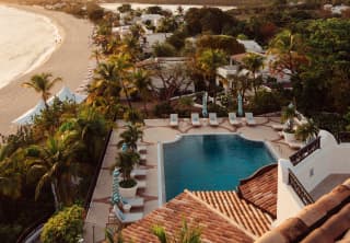 Sunset glows on the sand, roof tiles, palm trees and pool in a birds-eye view of the resort and distant sweep of Baie Longue.