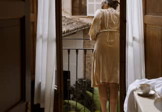 A woman in a gown leans over her balcony to see what's happening in the honey-stoned street below, viewed through open doors.
