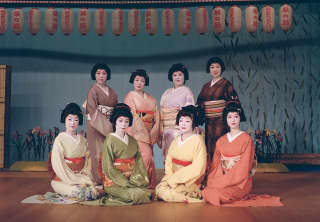 Eight women in traditional pastel Japanese kimonos and pale make-up pose below a row of lanterns, photographed by Cecy Young.