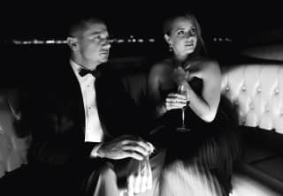 Black and white photograph of Jeremy Renner and Amy Adams, holding a glass of wine, as they sit in a luxurious water taxi.