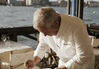 Chef Roberto Gatto, wearing chef's whites, dresses a plate of thinly-sliced bresaola at a Cip's Club waterside dining table.