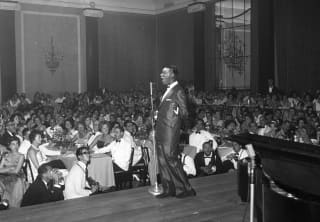 Black and white image of Nat King Cole in a dark suit, presenting on stage to a full audience in the Golden Room in 1959.