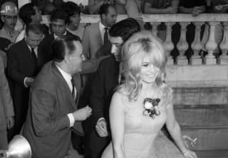 Seen from above, Brigitte Bardot, in a short dress with shaggy blond hair, smiles during a busy outside event at the hotel.