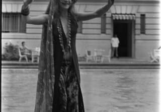 Singer Janis Joplin, wearing round glasses, head scarf, long beads, dress and cloak, puts her hands in the air by the pool.