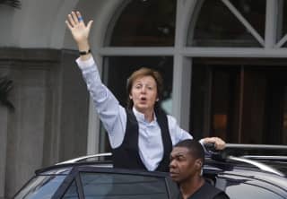 A guard stands by as Sir Paul McCartney in a blue shirt and black waistcoat waves from the open car door outside the hotel.