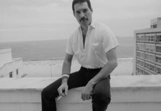 Black and white photo of Freddie Mercury sitting on a roof terrace wall in black pants and white shirt, holding a cigarette.