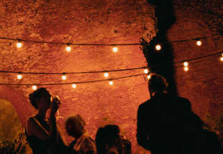 Mingling guests are silhouetted against the reddish glow of an ancient stone wall, illuminated by strings of bulb lights.