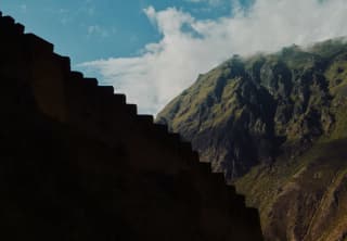 The steps of the Macha Picchu ruins, silhouetted in profile against the green-topped mountains and blue, cloudy sky, behind.