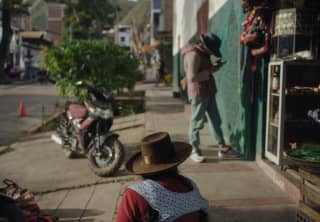 A man in a fedora checks his phone by a shop opening, watched by a local woman wearing a brown bolero hat, seen from behind.