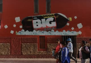 People walk past a building painted red with a mural of a bottle on its side on a layer of ice advertising Mega Big Cola.