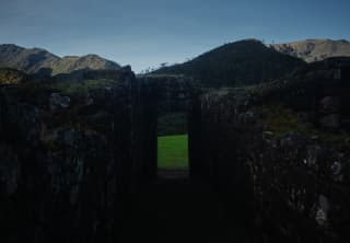 The doorway in the shadowy Llactapata stone ruins reveals a verdant grass area with a backdrop of mountainous ridges.