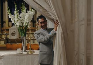 A man in a grey suit pulls drapes aside to reveal a clothed table with a vase of gladioli in the Palacio Nazarenas chapel.