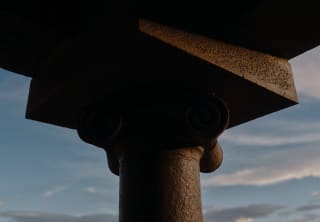 An ancient column's capital and architrave stands tall against a moody cloud-strewn sky of blue and grey, in close-up.