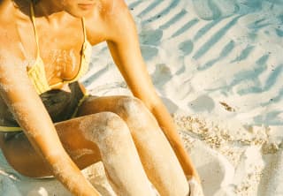 Close-up of a woman in a yellow bikini as she sits on the beach with her legs covered in sand, seen from above.