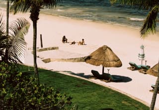 Three people relax on a white beach between palm trees and the sea, striped with shades of aquamarine, seen from above.