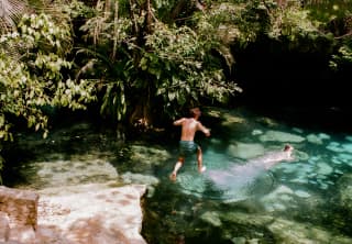 A man leaps of the pink rock edge of an open cenote where a friend already swims in the incredible turquoise waters.