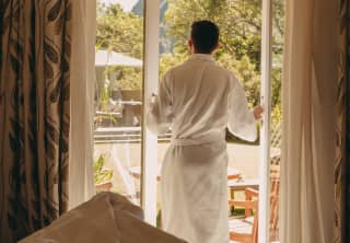 Seen across an unmade bed, a guest in a white bath robe opens the doors to his private terrace to greet the morning views.