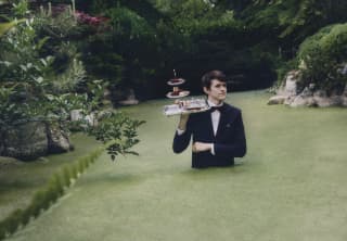 Surreal image of a waiter with a tray of afternoon tea cakes on his shoulder as he walks waist high through green pond weed.