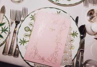 A place setting with cutlery and a plate with green botanical print topped with a pink Carriage Club menu, seen from above.