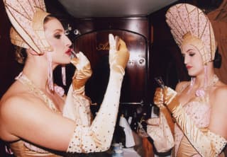 Two female Carriage Club performers, wearing flamboyant Art-Deco pink, sparkly headdresses apply red lipstick before a show.