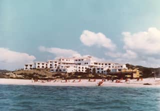 Historic image of beach-goers on the sand in front of Romazzino's hillside cluster of white buildings, seen from the sea.