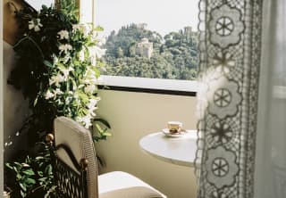 A coffee sits on a table in a warm terrace corner with a chair and blooming orange blossom, seen from behind a lace drape.