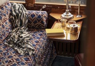 A lamp, a pot of tea and a book rest on a table in a cabin interior, with upholstered banquette in rich blues and maroons.