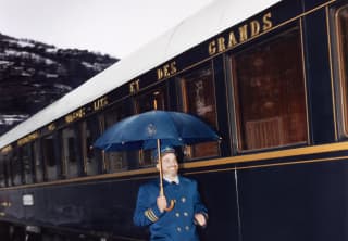 A steward in blue coat with gold buttons and cuff stripes smiles as he dashes through the rain below a matching blue umbrella