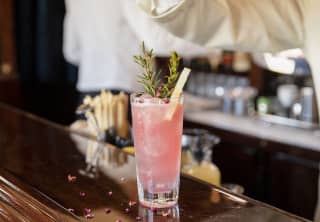 A barman's fingers scatter petal garnish on a grapefruit-pink cocktail with a rosemary sprig, on the gleaming wood bar top.