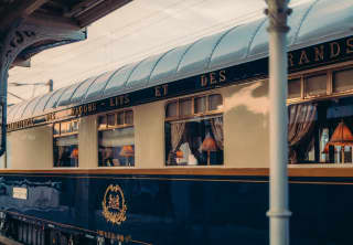 The cream, blue and gold livery of the dining car's exterior shines in the early evening sun. Inside table lamps glow orange