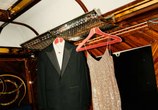 A sparkling gold evening dress and a tuxedo dangle from coat hangers above a table and a blue velvet chair in a cabin.