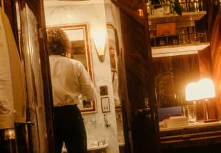 A man in black trousers and white shirt checks his tie in the bathroom mirror, seen from the warm-lit cabin interior behind.