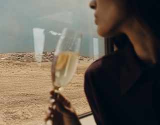 In soft-focus foreground, a seated woman holds her glass of sparkling wine at a window table as sand-hued plains pass by.
