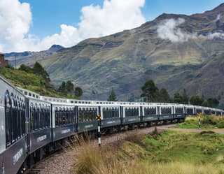 Blue and white train carriages curving under a cloud-coated Andean mountain