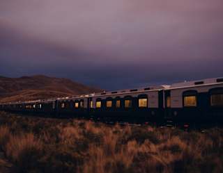 Lights glow in cabin windows as the train journeys through the evening beneath a purpling sky, seen from grasslands outside.