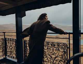 A guest in a long brown coat leans on the observation deck's filigree rails gazing out over gold grasslands and a blue lake.