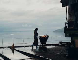 A woman stands by a raised firepit on a platform overlooking Lake Titicaca, silhouetted against grey sky and silvery water.