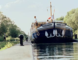 Water-level view of the Amaryllis on a narrow section of the Canal du Centre, about to be moored by a man holding a rope.