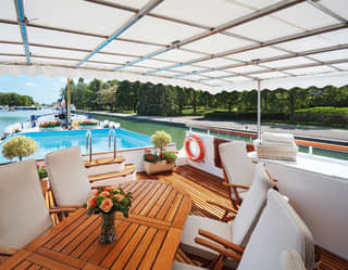 Wooden table surrounded by white fabric chairs on the top deck of a luxury barge