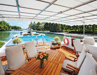 Wooden table surrounded by white fabric chairs on the top deck of a luxury barge