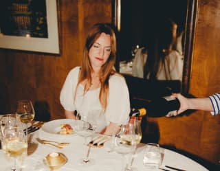 At a dining table, a female guest watches as white wine is decanted into her glass, before tasting her elegant starter dish.