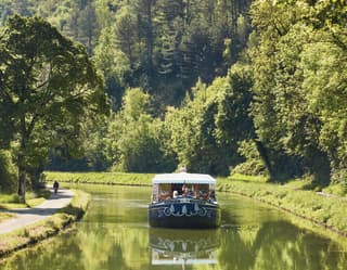 The Fleur de Lys cruises up the serene Ouche river, bordered by big trees and green banks, where a cyclist enjoys the route.