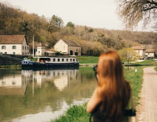 A woman stands on the bank of the canal, taking a photograph of the Fleur de Lys as it cruises by a cluster of white houses.