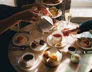 A woman pours a large cup of tea for a friend at a table with a tiered-stand of sweet and savoury bites, with scones and jam.