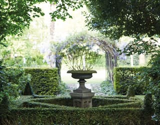 Low topiary box hedges form a diamond shape around a stone bird bath in lush gardens fills with mature trees