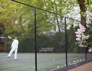 A man in white finishes a tennis shot on a green hard court. Behind him multiple yellow balls line the base of the high fence