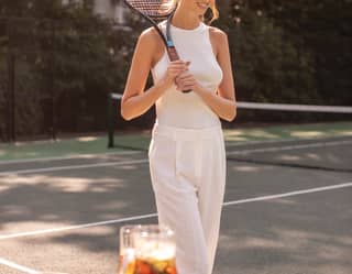A woman in white rests a racquet on her shoulder. Before her is a table with a jug of Pimms, plus a spare racquet and ball